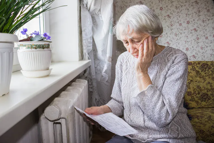 An elderly woman reads a bill by the radiator with despair.