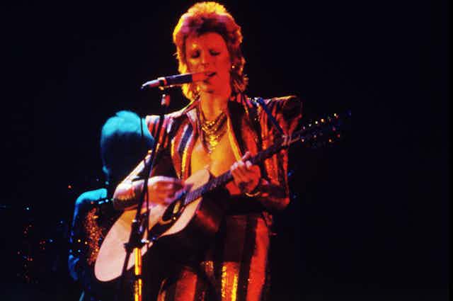 Hombre cantando y tocando la guitarra con un atuendo glam-rock de los años 70.