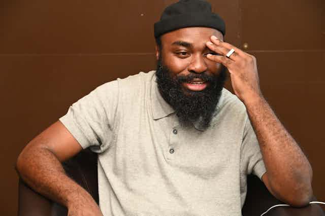 A bearded man in a beanie and T-shirt smiles as he expresses himself, the fingers of one hand placed on his forehead.