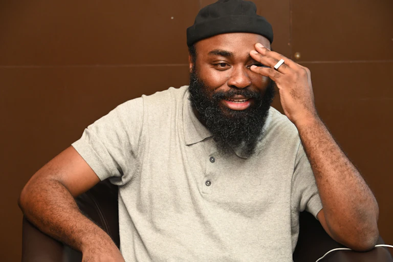 A bearded man in a beanie and T-shirt smiles as he expresses himself, the fingers of one hand placed on his forehead.