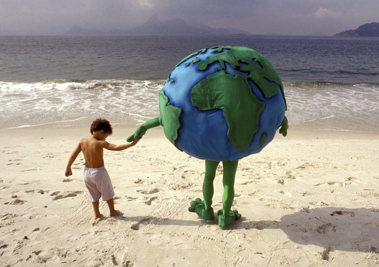 A person in a costume of the Earth holds a child's hand on a beach in Rio. The photo is from 1992