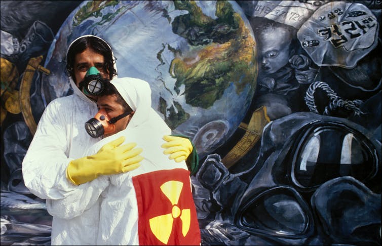 A young man holding a nuclear symbol on a contamination suit embraces another man wearing a gas mask in front of a dark depiction of Earth.