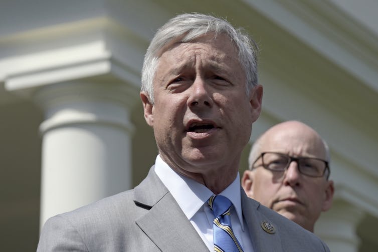 A man with gray hair, a gray jacket, white shirt and blue tie talking outside a building.