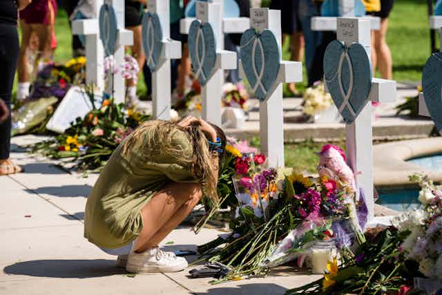 A woman squats on the floor next to piles of flowers, and memorials.