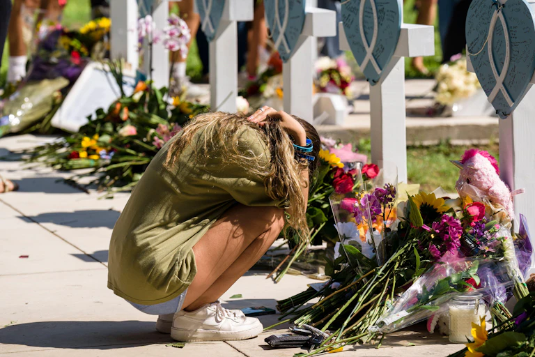A woman squats on the floor next to piles of flowers, and memorials.