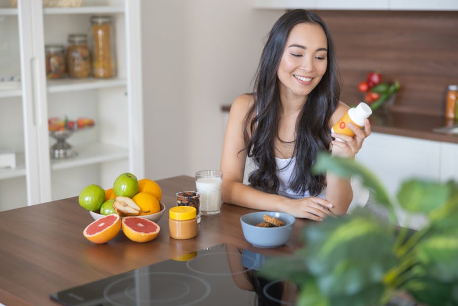 A young woman looks at a bottle of vitamin supplements in her kitchen, next to a bowl of fruit.