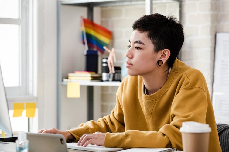 An Asian woman wearing a yellow sweatshirt and black tunnel earrings sits at a computer with an LGBTQ+ flag in the background.