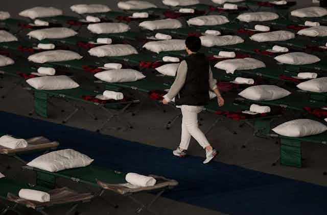 A woman in white pants and a black vest walks past dozens of cots with pillows, blankets and towels on them.