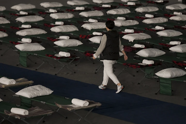 A woman in white pants and a black vest walks past dozens of cots with pillows, blankets and towels on them.