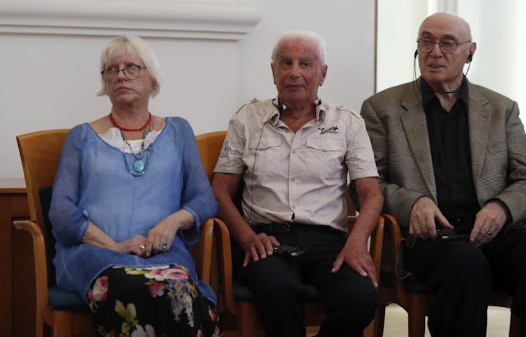 An elderly woman in a blue shirt sits next to two elderly men.