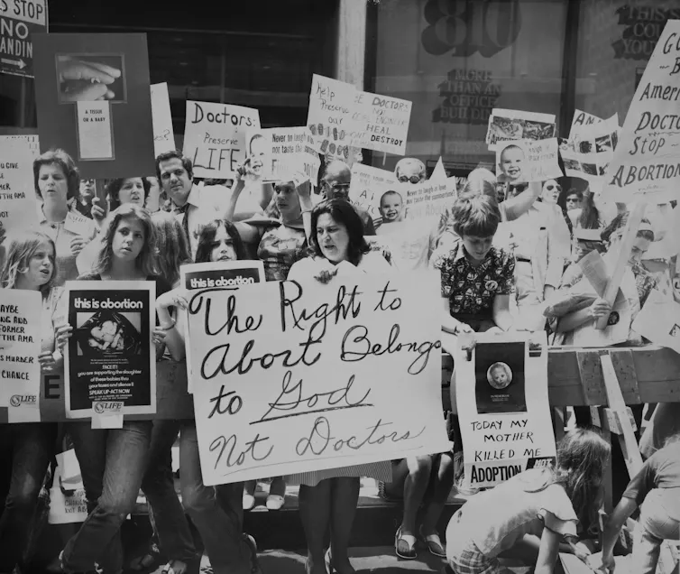 A black and white photo shows demonstrators with signs that say things like 'the right to abortion belongs to God, not doctors.'