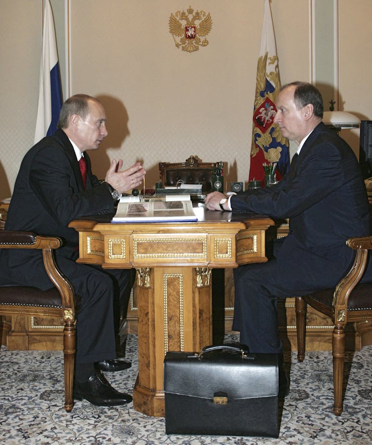 Two white men dressed in business suits sit across from each other at desk.