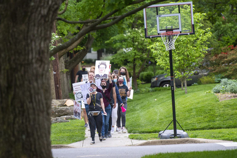 Women march with a megaphone and a hanger and a photo of Kavanaugh that says liar, in a suburban street.