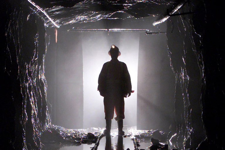 A man stands silhouetted through a door way in a mine shaft, he's wearing a hard hat
