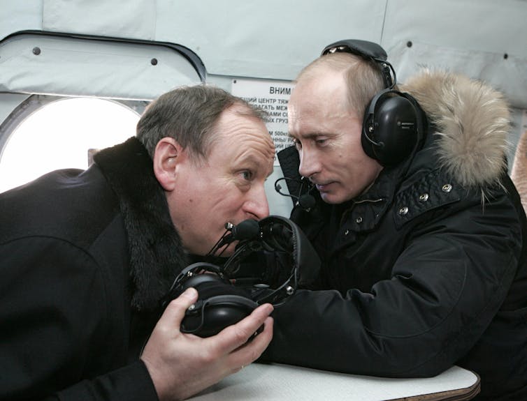 One man is talking while another man holds his ear muffs and is listening.