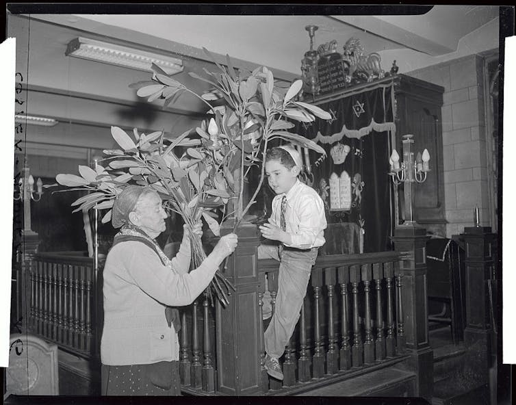 In this black and white photo, an elderly woman and young boy tie tree branches to a post as decoration.