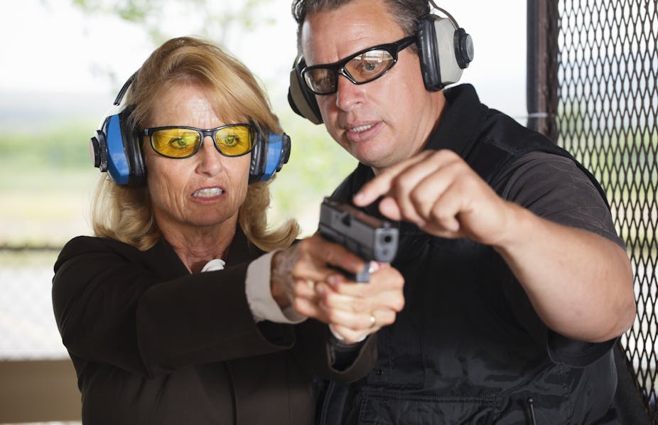 A male firearms instructor stands alongside a woman firing a gun at a gun range.