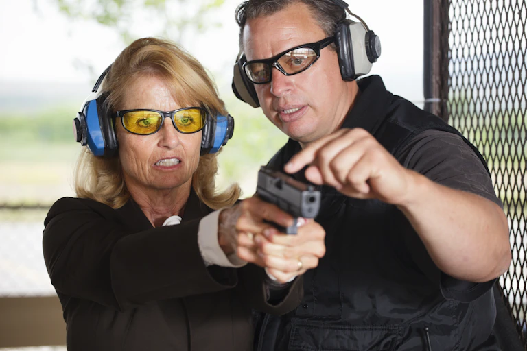 A male firearms instructor stands alongside a woman firing a gun at a gun range.