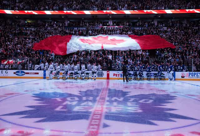 A hockey rink is seen in the foreground with players standing with a massive Canada flag over the crowd in the background