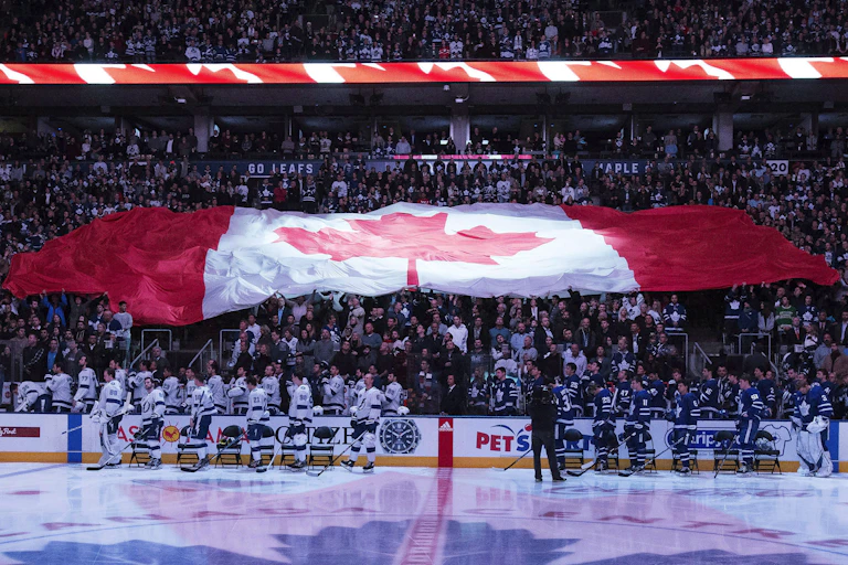 A hockey rink is seen in the foreground with players standing with a massive Canada flag over the crowd in the background