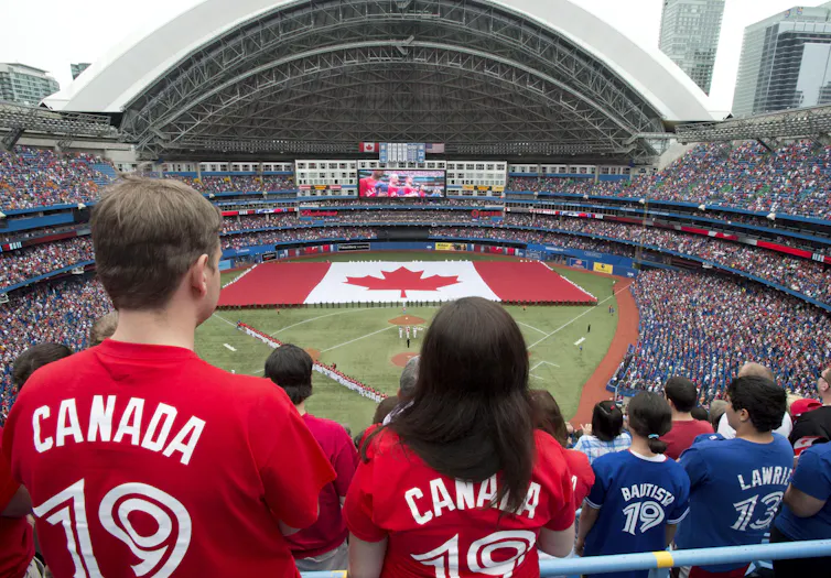A man and a woman with blue jays jerseys that say canada on the back stand with the field in the background