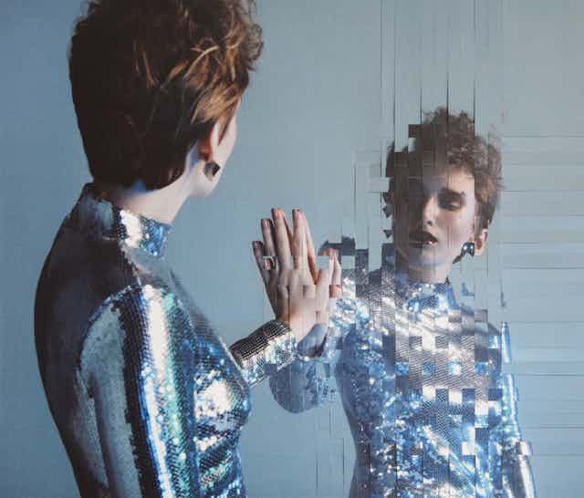 A woman wearing a shiny metallic top presses her hand against a mirror that appears to be in a woven pattern of inch-long strips.