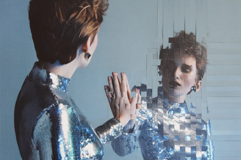 A woman wearing a shiny metallic top presses her hand against a mirror that appears to be in a woven pattern of inch-long strips.