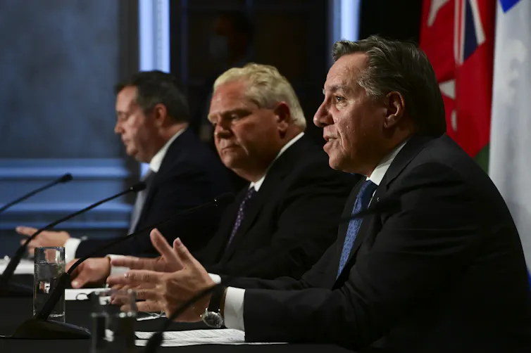 Three men in suits seen seated at a press conference in front of microphones.