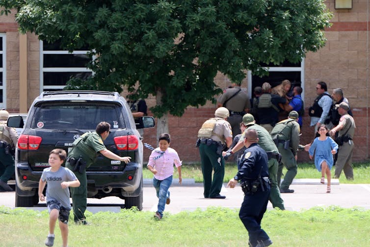 Young children are seen being led out of a classroom window and sprinting away from a school while police officers point where they should be going.