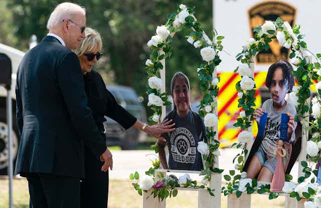 Joe Biden, a grey-haired man in a suit, and his wife Jill Biden, a woman in a suit, look reverently at images of schoolchildren surrounded by flowers, a memorial to the victims of the Texas school shooting.