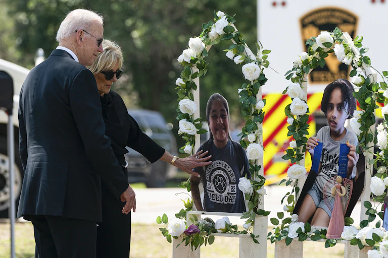 Joe Biden, a grey-haired man in a suit, and his wife Jill Biden, a woman in a suit, look reverently at images of schoolchildren surrounded by flowers, a memorial to the victims of the Texas school shooting.