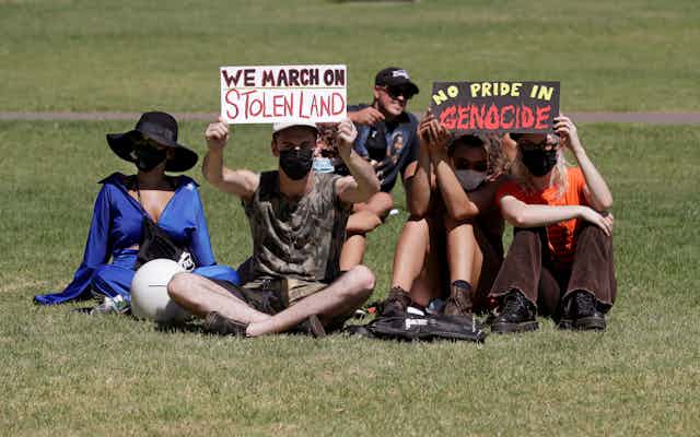 People sit in the sun holding signs saying 'we march on stolen land' and 'no pride in genocide'.
