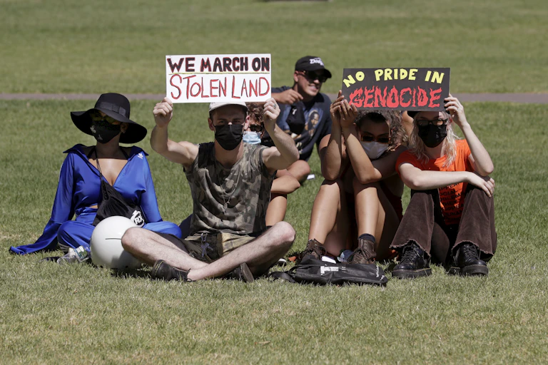 People sit in the sun holding signs saying 'we march on stolen land' and 'no pride in genocide'.