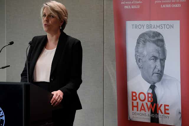 Woman speaking at a podium in front of poster promoting biography of Bob Hawke