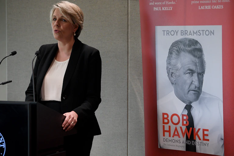 Woman speaking at a podium in front of poster promoting biography of Bob Hawke