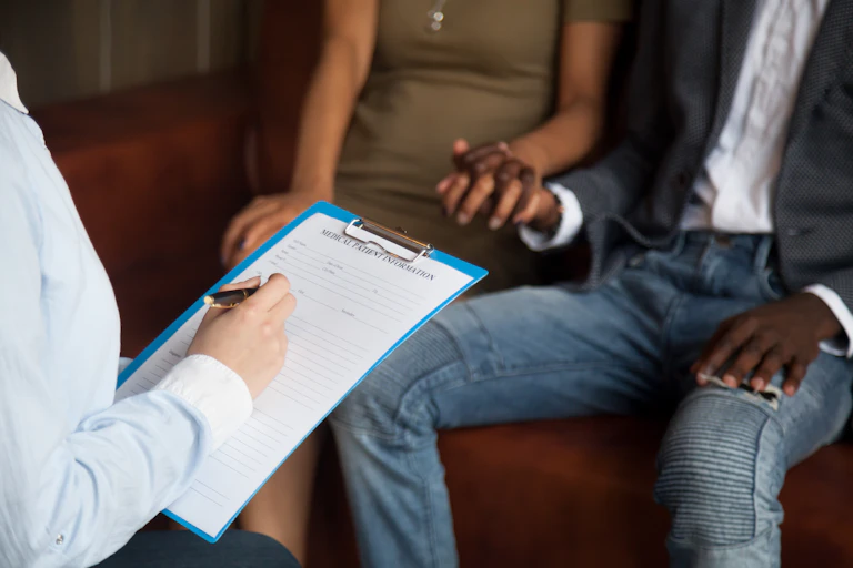 A heterosexual couple holds hands while they're doctor fills out a patient information form on a clipboard.