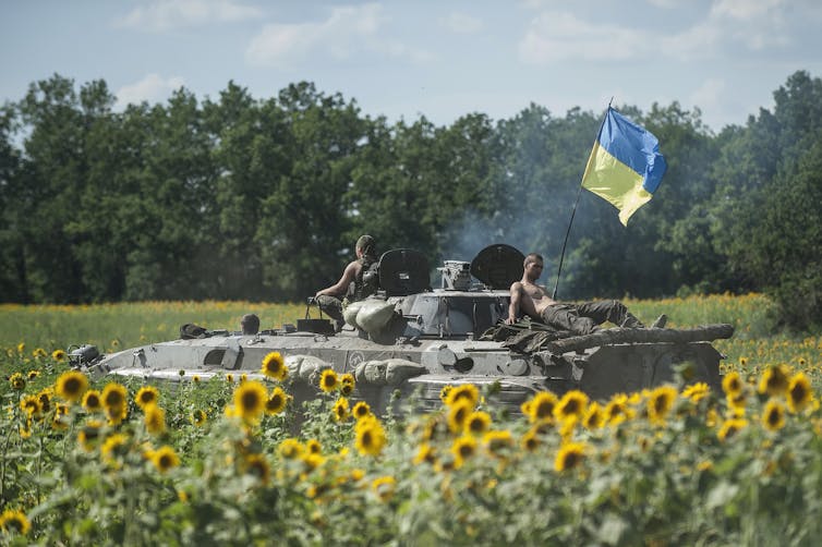 Bare-chested soldiers sit in the sun atop a military vehicle adorned with a Ukrainian flag. Sunflowers are in the foreground.