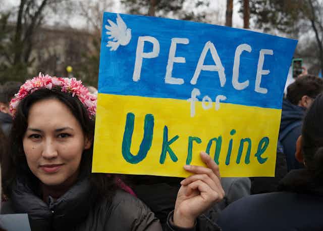 A dark haired woman wearing a pink flower garland in her hair holds up a blue and yellow sign that reads Peace for Ukraine.
