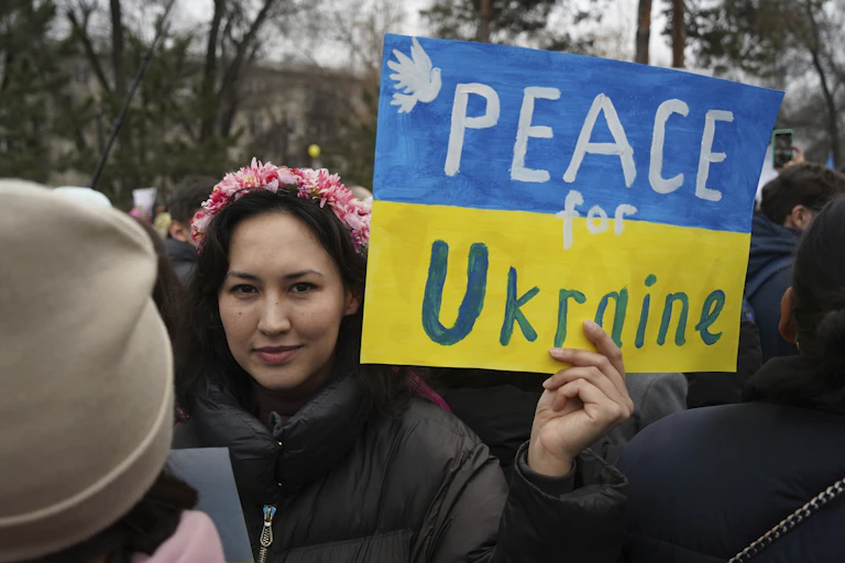 A dark haired woman wearing a pink flower garland in her hair holds up a blue and yellow sign that reads Peace for Ukraine.