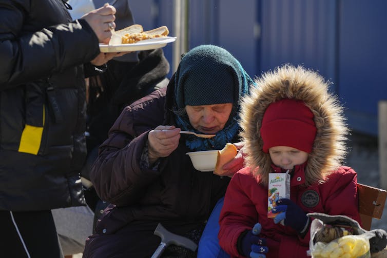 An elderly woman eats soup while a boy in a red parka sips on a juice box.