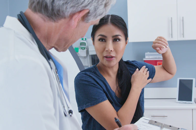 A woman holding her left arm up and pointing to her left shoulder with her other hand, speaking to a man in a white coat with at stethoscope seen from behind