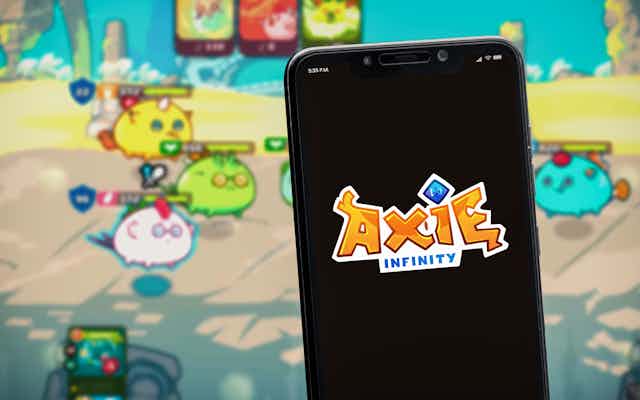 A mobile phone with the Axie Infinity logo is held up in front of a screen with the game being played on it. 