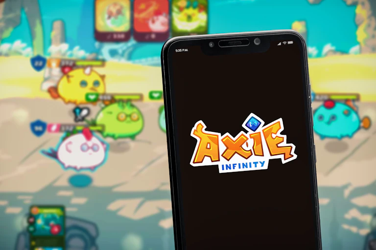 A mobile phone with the Axie Infinity logo is held up in front of a screen with the game being played on it.