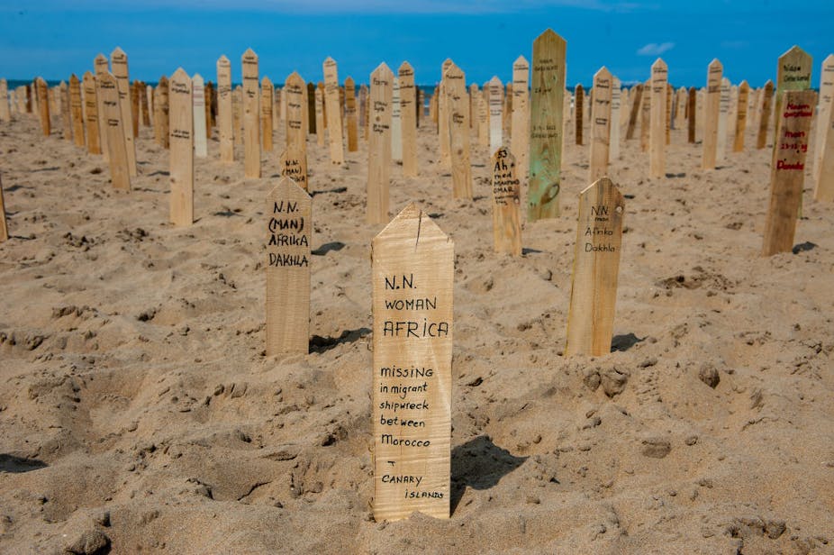 Planted in beach sand are wooden spikes, each with a name and some details about them.