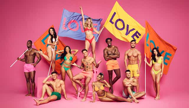 The cast of Love Island, 11 attractive young people in neon bikinis and swim trunks, posing with large colourful flags reading LOVE, against a bright pink background
