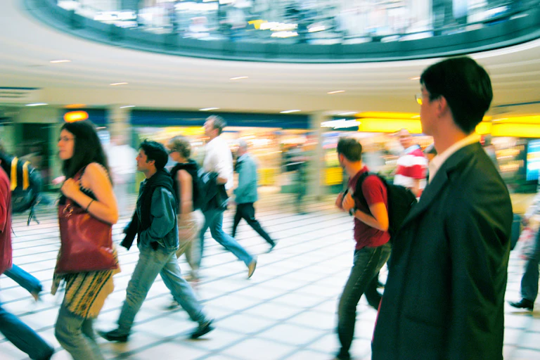 Commuters walk fast through a brightly lit rail station hall.