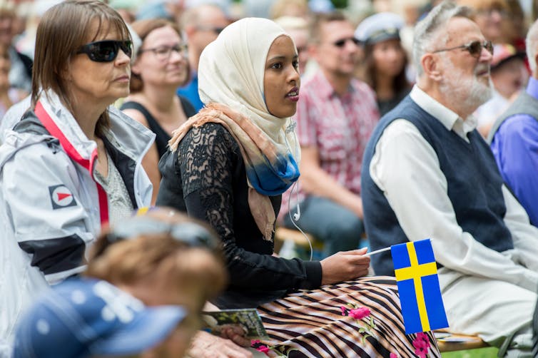 Men and women sit in a crowd holding Swedish flags.