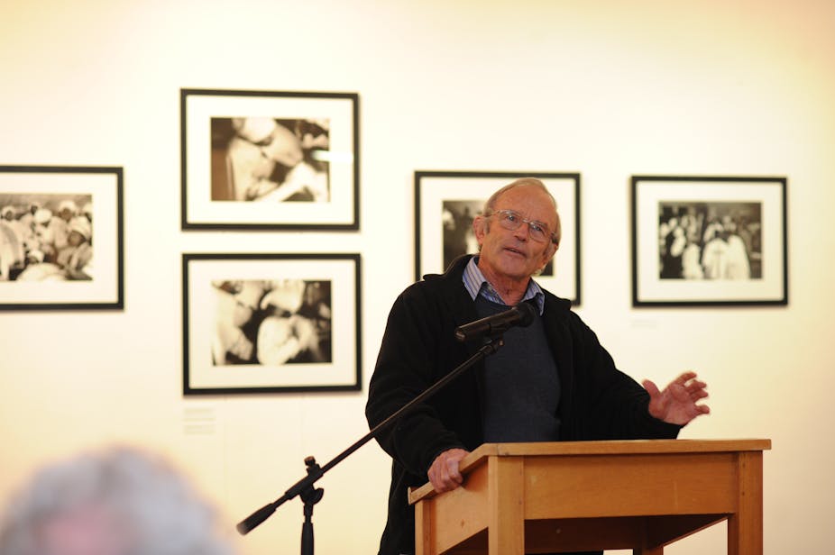 An elderly man in glasses stands talking into a microphone in a room with framed black and white photos behind him.