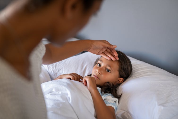 A young girl lies in bed. An adult feels her forehead.