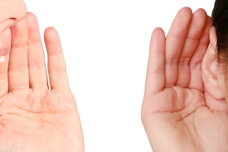 Close up of faces of two people, one cupping her hand around her mouth and speaking, the other cupping hand around her ear and listening
